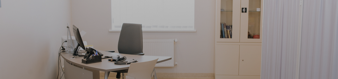 Minimal medical clinic consultation room with desk, chair, computer and storage cabinets, representing a professional cosmetic or healthcare setting.